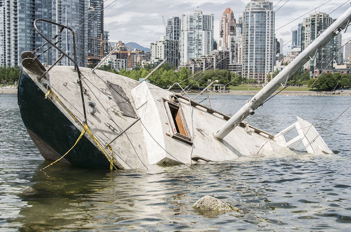 A partially submerged white sailboat is pictured in murky green water with apartment buildings in the background.