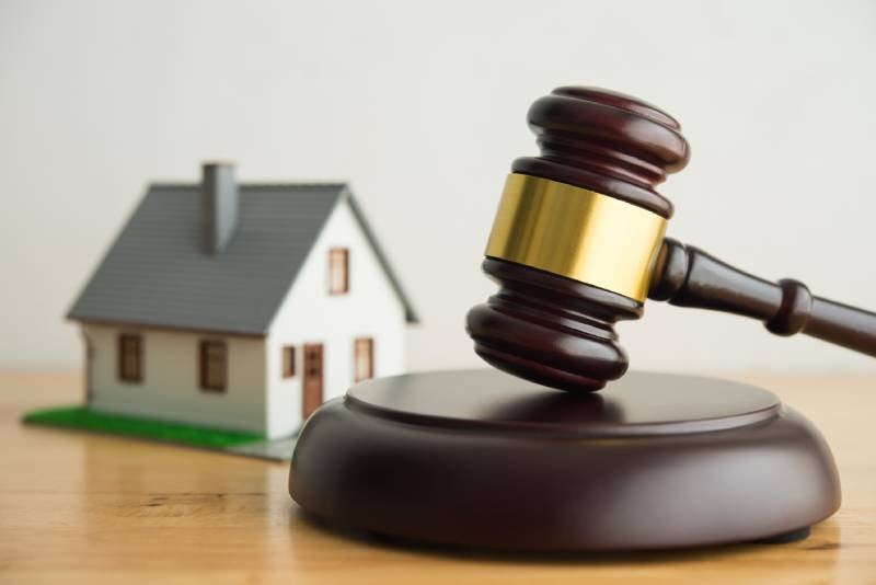 A wooden gavel rests on a sounding block, with a blurred miniature house in the background.